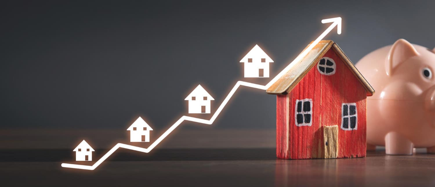 A wooden toy house next to a piggy bank, with homes and a rising graph line symbolizing a growing housing market.