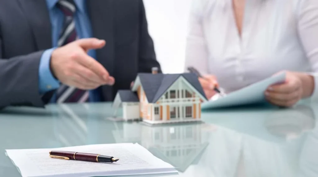A businessman gestures while discussing real estate details with a woman, who takes notes, beside a model house and paperwork.