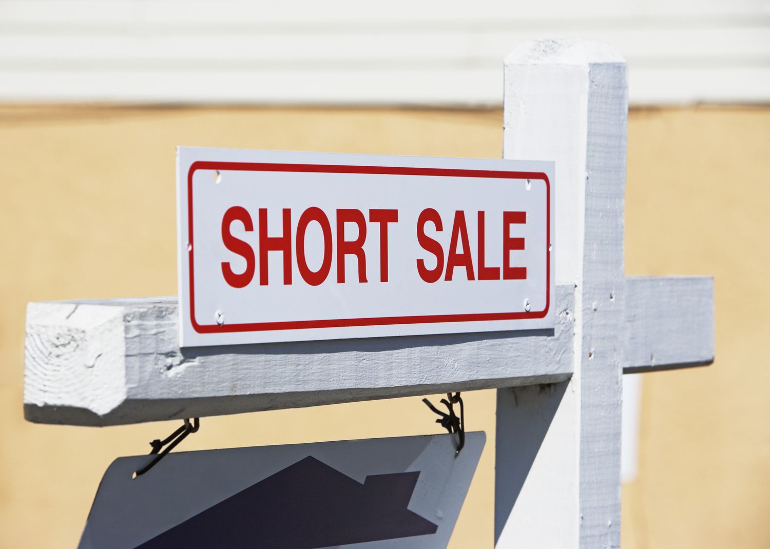 A real estate sign reading "SHORT SALE" in bold red letters, attached to a white wooden post against a beige background.