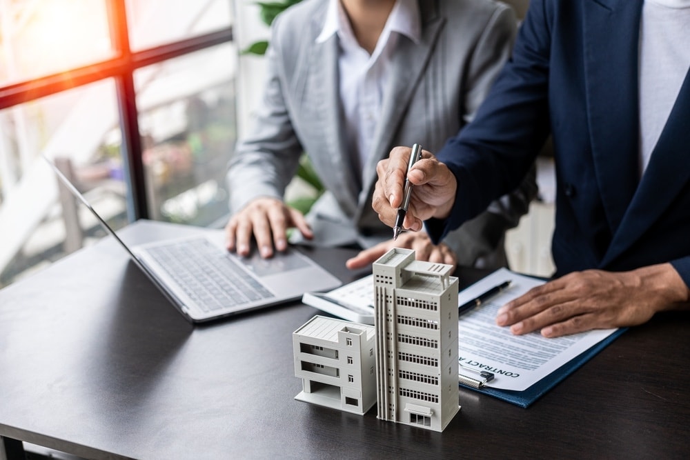 Two business professionals signing a contract beside a laptop and a miniature building model on a desk. Bright windows in background.