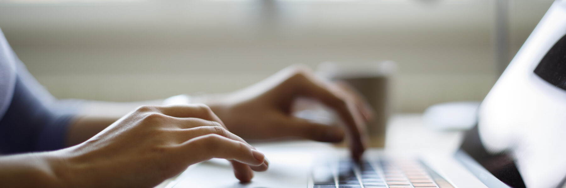 Close-up of a person’s hands typing on a laptop keyboard, perhaps brainstorming 25 summer social media ideas for real estate agents, with a blurred background featuring a coffee cup and sunlight streaming through the window.